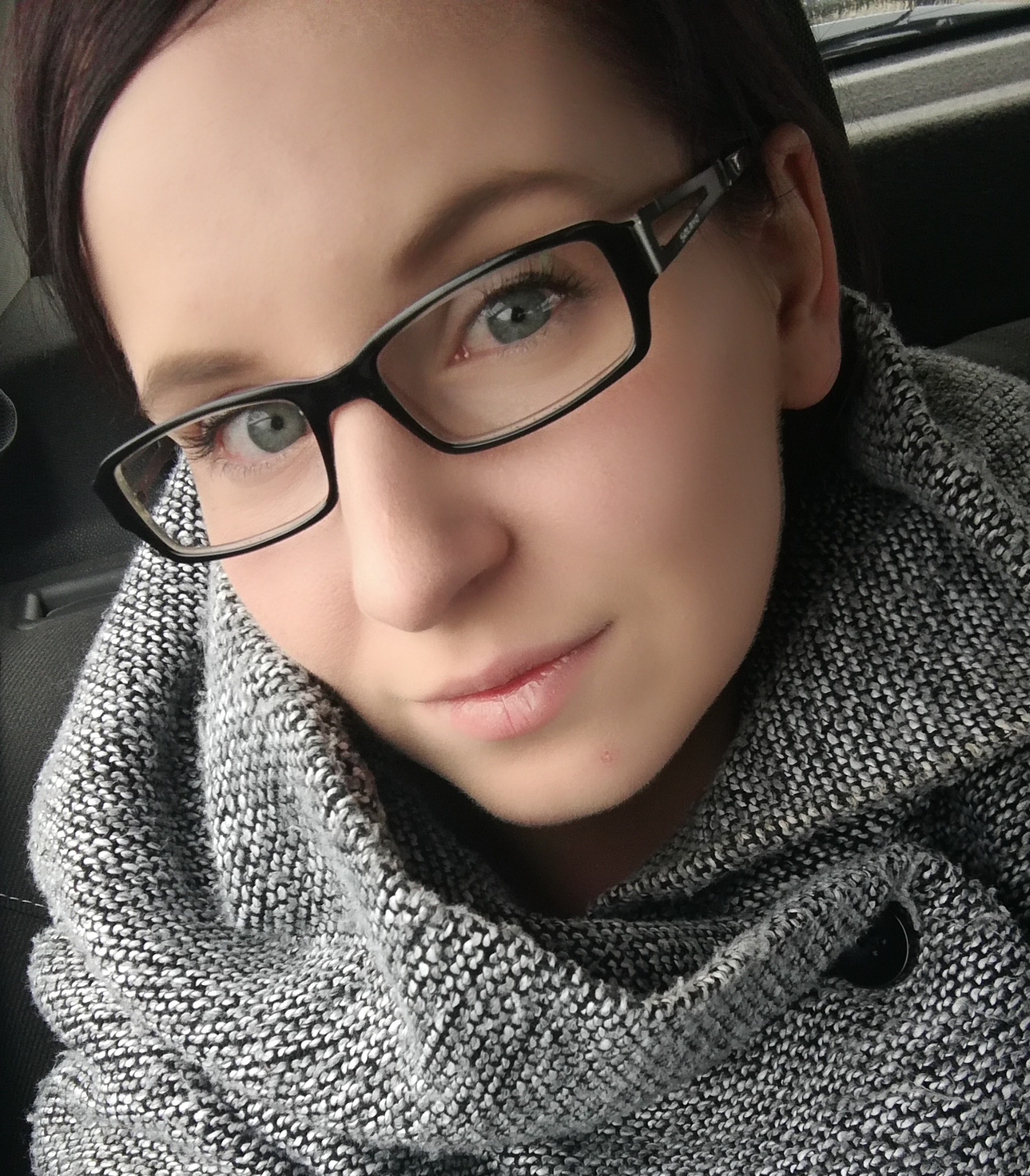 Portret kobiety w okularach i szarym, dzierganym swetrze z dużym kołnierzem, czarne oprawki okularów z widocznym logo na zauszniku.