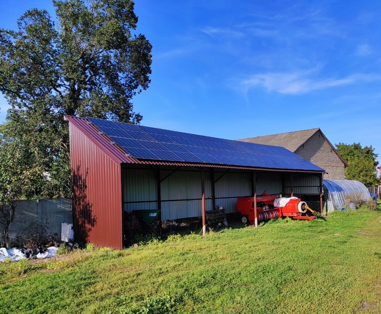 Instalacja fotowoltaiczna na zadaszeniu w gospodarstwie rolnym, panele w kolorze ciemnoniebieskim, w tle maszyna rolnicza i budynek gospodarczy.