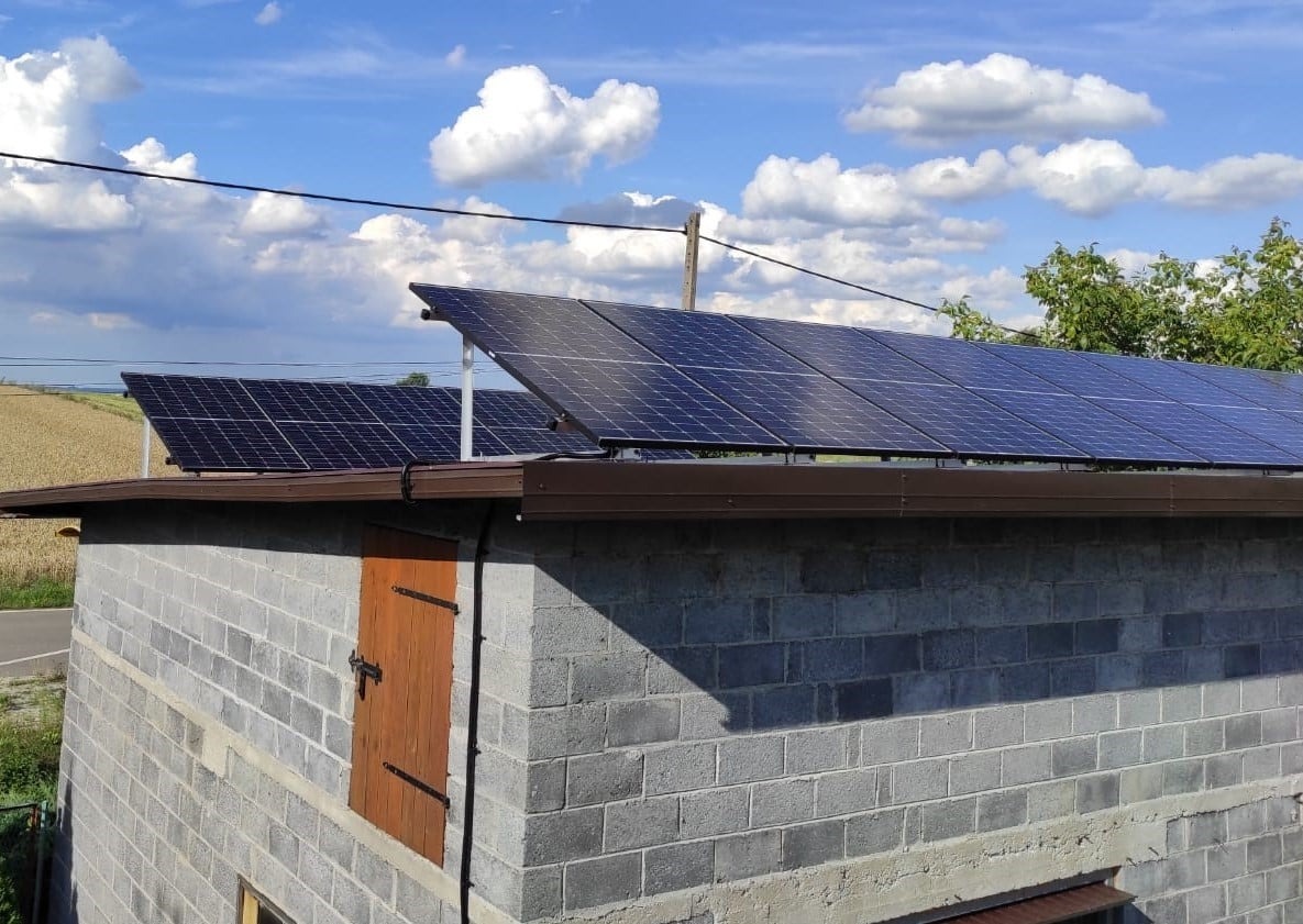 Instalacja paneli słonecznych na dachu budynku gospodarczego z szarej cegły, widok na pola i błękitne niebo z chmurami.