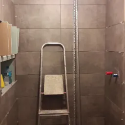 Układanie brązowych płytek w łazience, widoczna drabina, listwa wykończeniowa i wentylator, etap prac wykończeniowych.