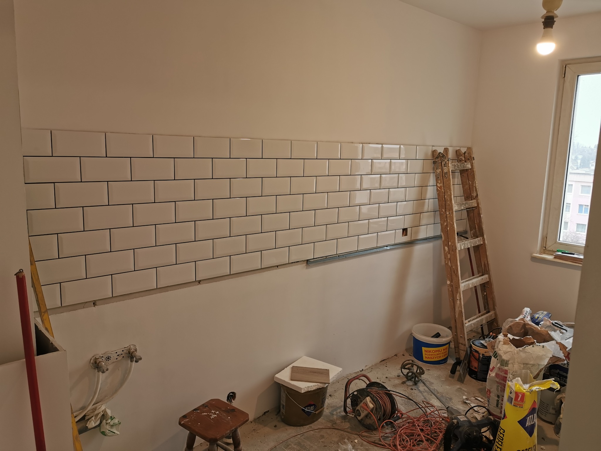 Fragment ściany z ułożonym rzędem białych, prostokątnych płytek, widoczne narzędzia, drabina i materiały budowlane, stanowiące wyposażenie wnętrza w trakcie prac wykończeniowych.