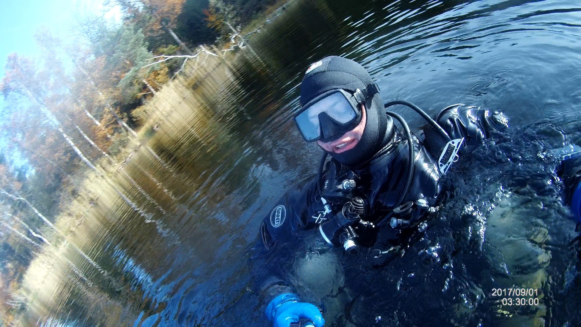 Nurek w pełnym ekwipunku zanurzony w jeziorze, widoczne odbicie drzew w wodzie i datownik w prawym dolnym rogu.