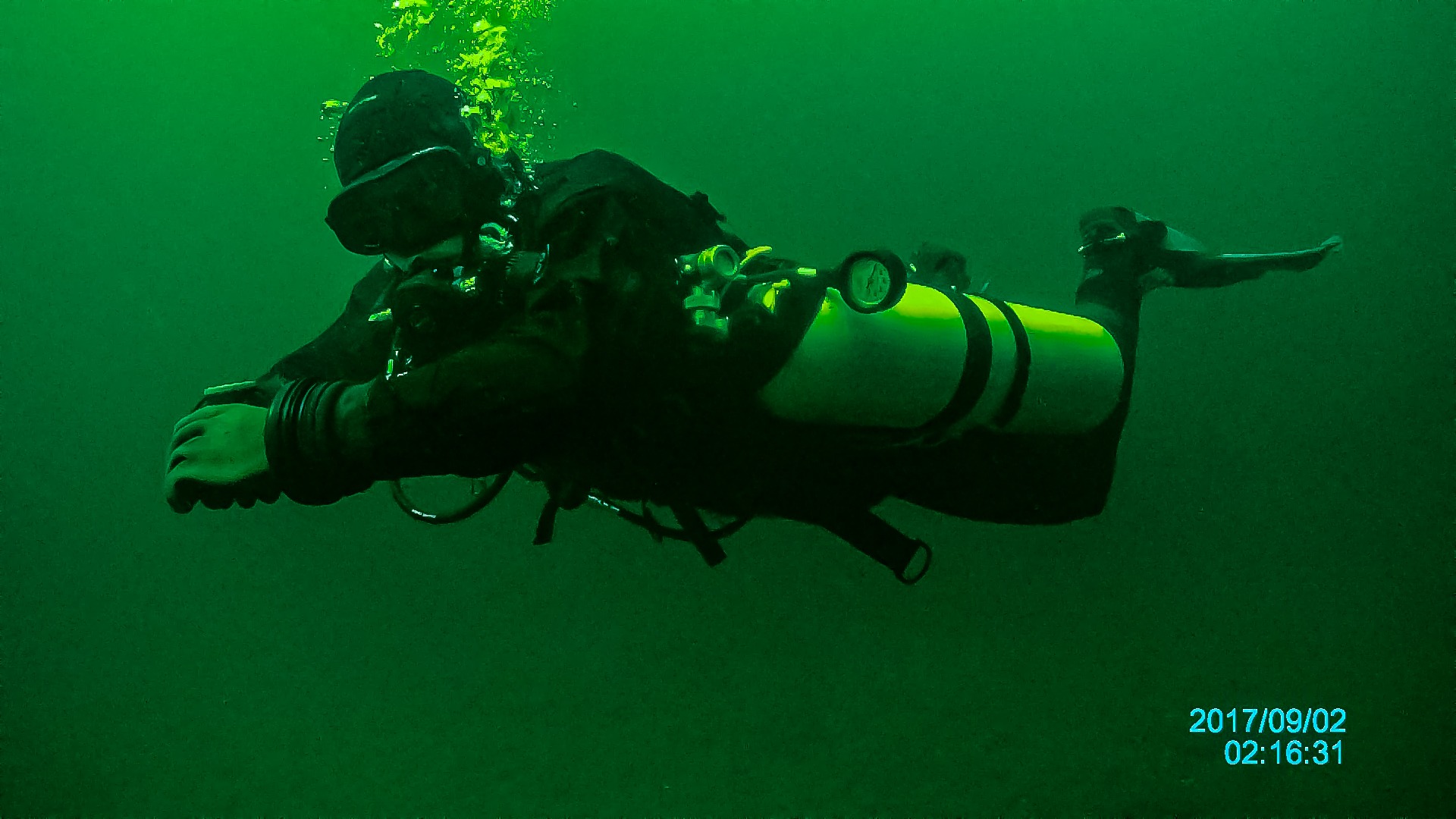 Nurek w pełnym ekwipunku, zanurzony w ciemnozielonej wodzie, widoczne bąbelki powietrza unoszące się ku powierzchni, data i godzina w prawym dolnym rogu.