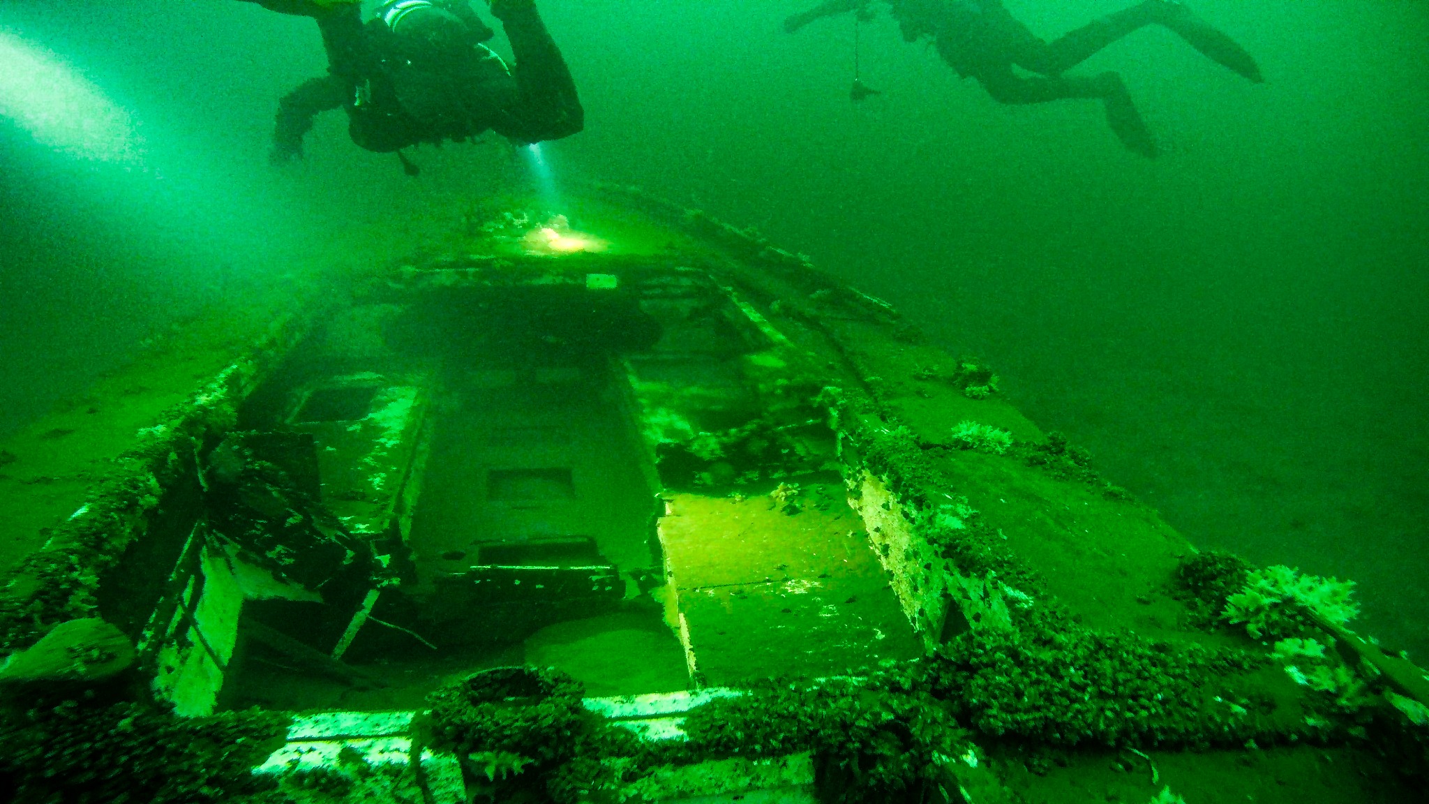 Zatopiony wrak pojazdu pod wodą, oświetlony latarką nurka, z dwoma nurkami w tle, widoczne porastające wrak rośliny.