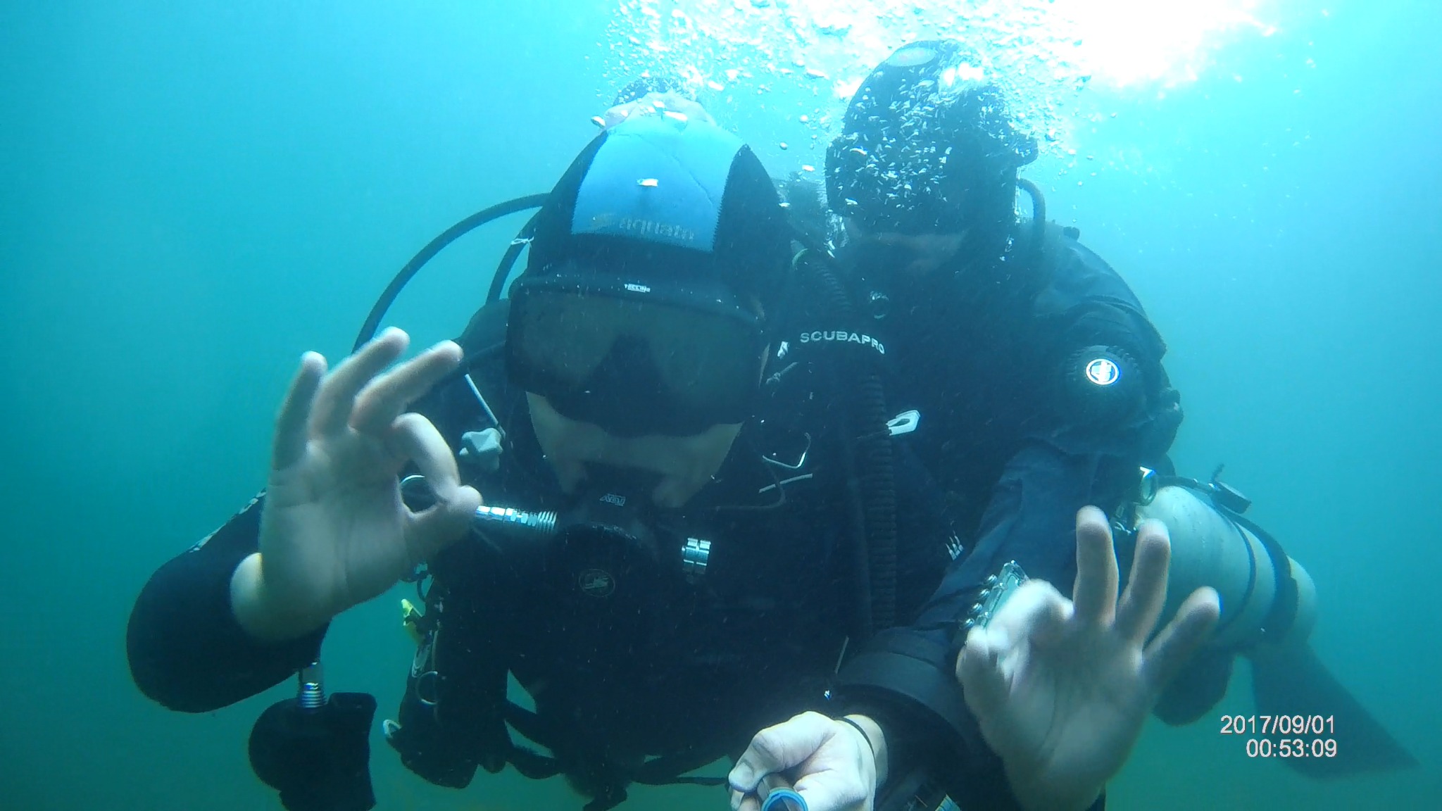 Dwóch nurków pod wodą, jeden pokazuje znak OK, widoczne maski, butle z tlenem, kombinezony, lekko zamulona woda.