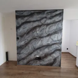 Ściana akcentująca w salonie w Krakowie z dekoracyjnym malowaniem imitującym marmur, widoczne gniazdka elektryczne i drewniana podłoga, w tle białe ściany i drzwi.