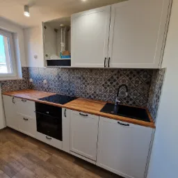 Nowoczesna kuchnia z białymi szafkami, drewnianym blatem i wzorzystą mozaiką na ścianie, widok na czarny zlew i płytę indukcyjną, oraz otwartą szafkę wiszącą z widoczną rurą wentylacyjną.