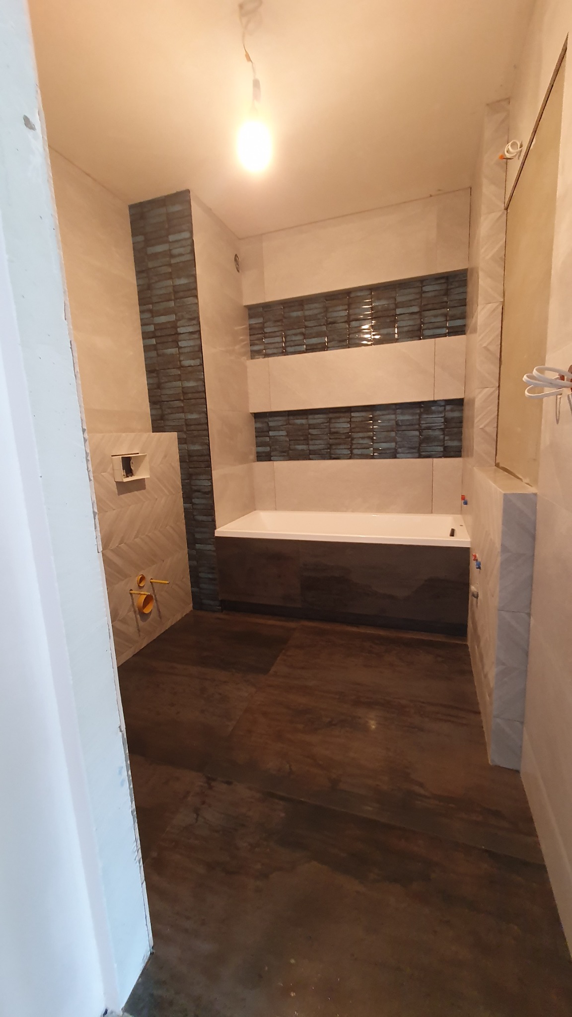 Niedokończona łazienka z wanną obudowaną ciemnym drewnem, mozaiką na ścianach i instalacją podtynkową WC z wystającymi rurami.