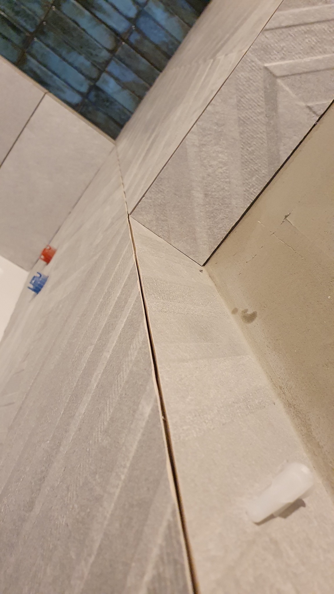 Układanie szarych płytek z delikatnym wzorem w jodełkę, widoczne krzyżyki dystansowe w kolorze niebieskim i czerwonym oraz biały klin poziomujący, fragment niebieskiej mozaiki w tle.