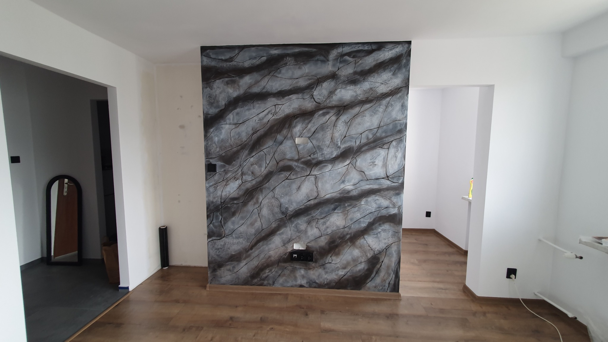 Ściana akcentująca w salonie w Krakowie z dekoracyjnym malowaniem imitującym marmur, widoczne gniazdka elektryczne i drewniana podłoga, w tle białe ściany i drzwi.