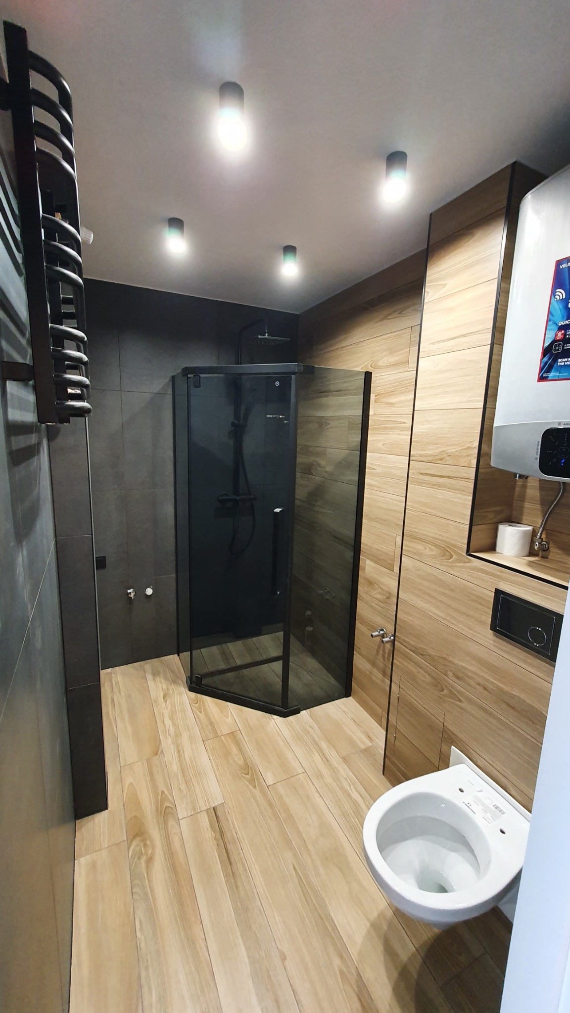 Nowoczesna łazienka z kabiną prysznicową typu walk-in z czarnymi ramami, drewnopodobnymi płytkami na podłodze i ścianie, szarymi płytkami na ścianie z grzejnikiem drabinkowym i podwieszaną toaletą.