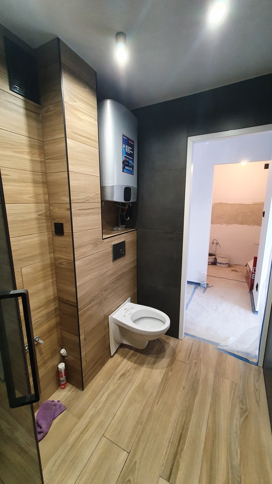 Nowoczesna łazienka w trakcie wykańczania, z drewnopodobnymi płytkami na podłodze i ścianach, podwieszana toaleta, bojler elektryczny, widok na sąsiednie pomieszczenie w stanie surowym.