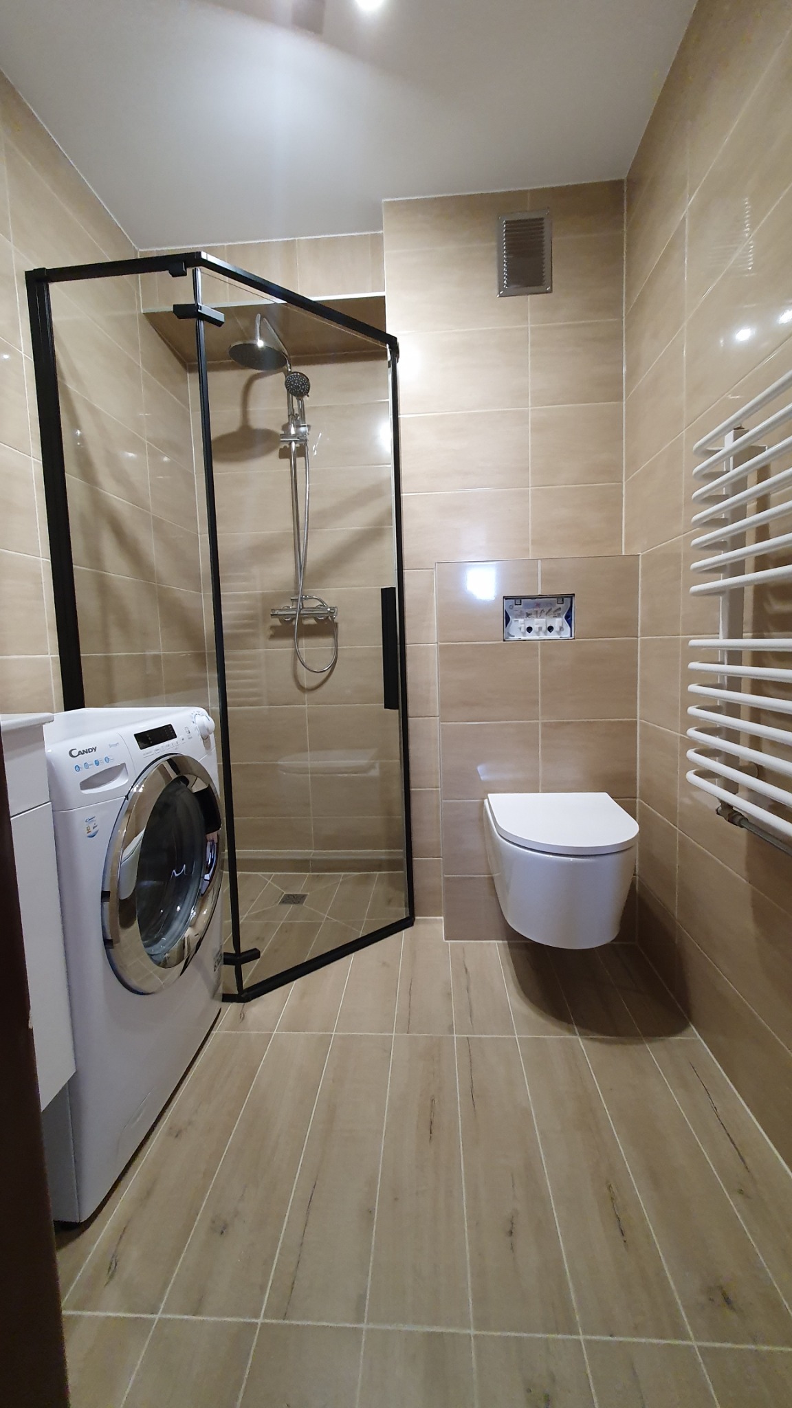 Nowoczesna łazienka z beżowymi płytkami, kabiną prysznicową typu walk-in z czarną ramą, podwieszaną toaletą, grzejnikiem drabinkowym i pralką Candy.