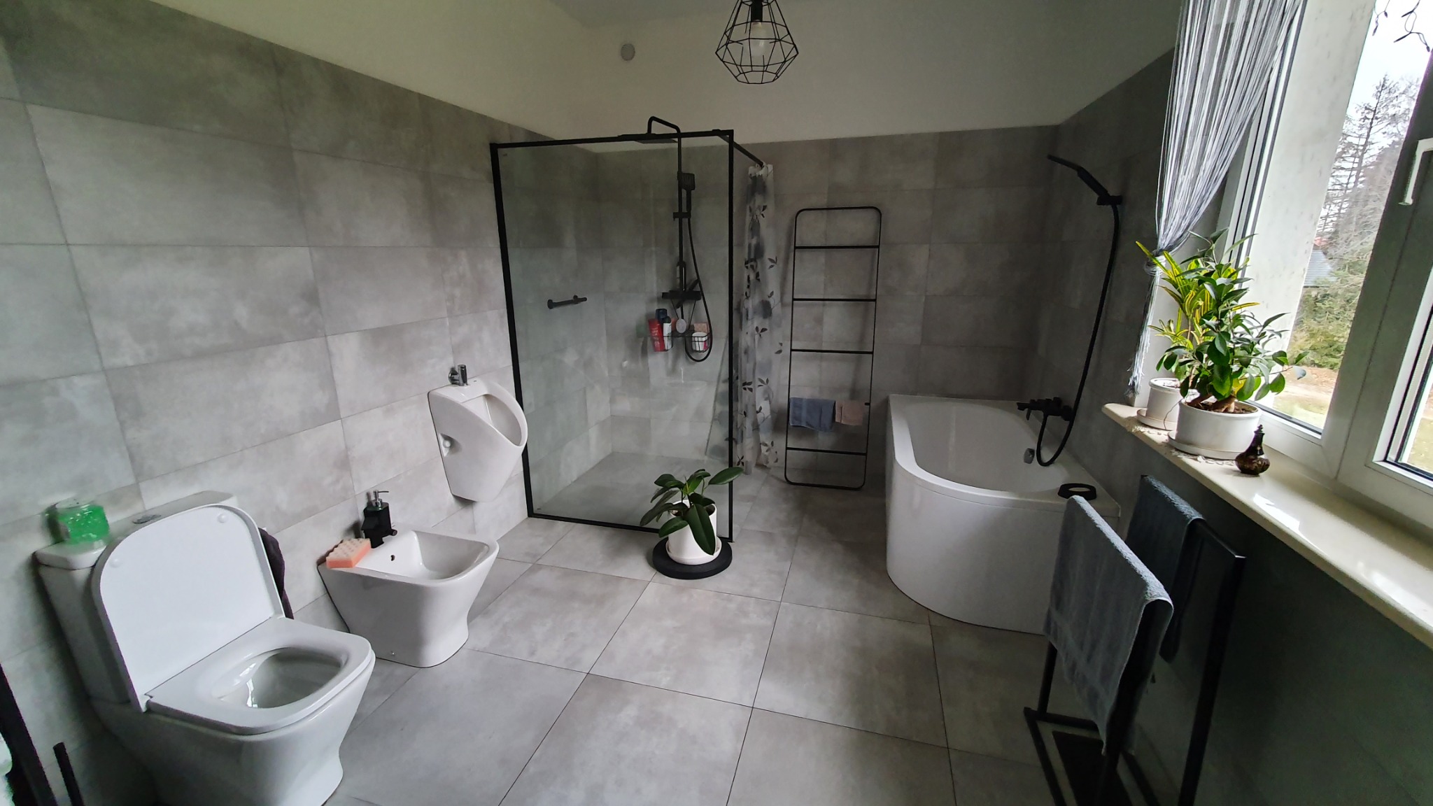 Nowoczesna łazienka w szarościach z kabiną prysznicową typu walk-in, wiszącym pisuarzem, toaletą, bidetem, wanną i roślinami doniczkowymi przy oknie.