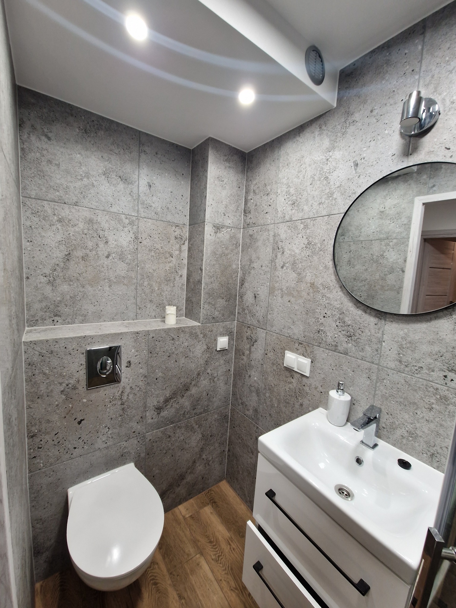 Nowoczesna łazienka z szarymi płytkami imitującymi kamień, białym wiszącym WC, białą szafką pod umywalką z czarnymi uchwytami oraz okrągłym lustrem z widocznym odbiciem fragmentu mieszkania.
