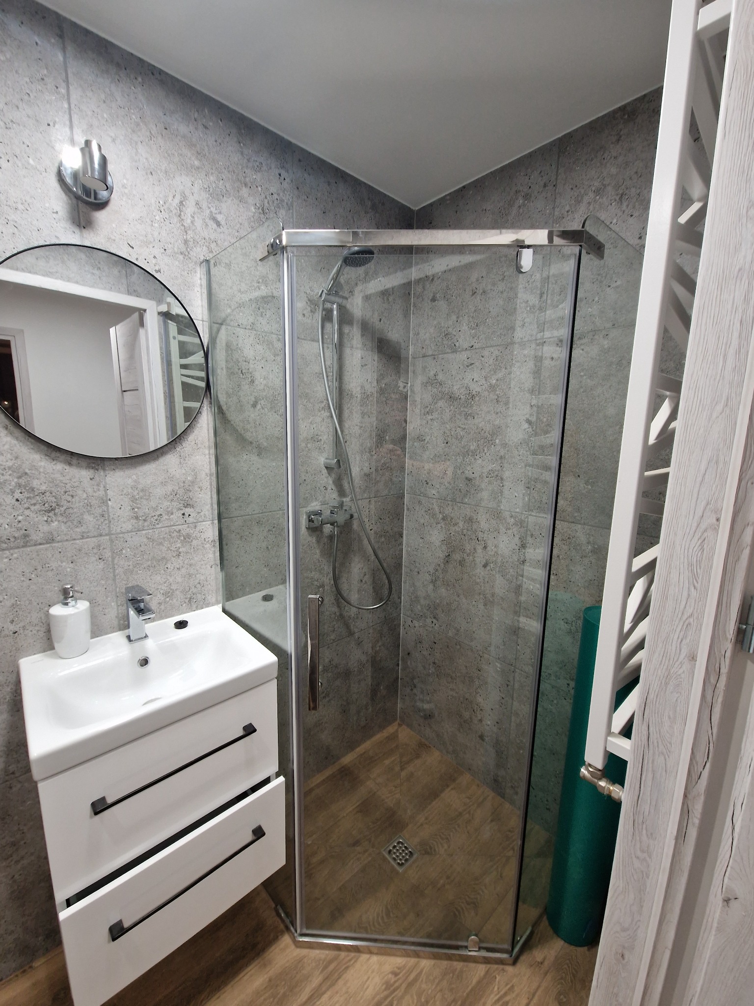 Nowoczesna łazienka z kabiną prysznicową, umywalką nablatową z szafką z szufladami z czarnymi uchwytami, okrągłe lustro z podświetleniem i grzejnik drabinkowy z drewnianym wykończeniem