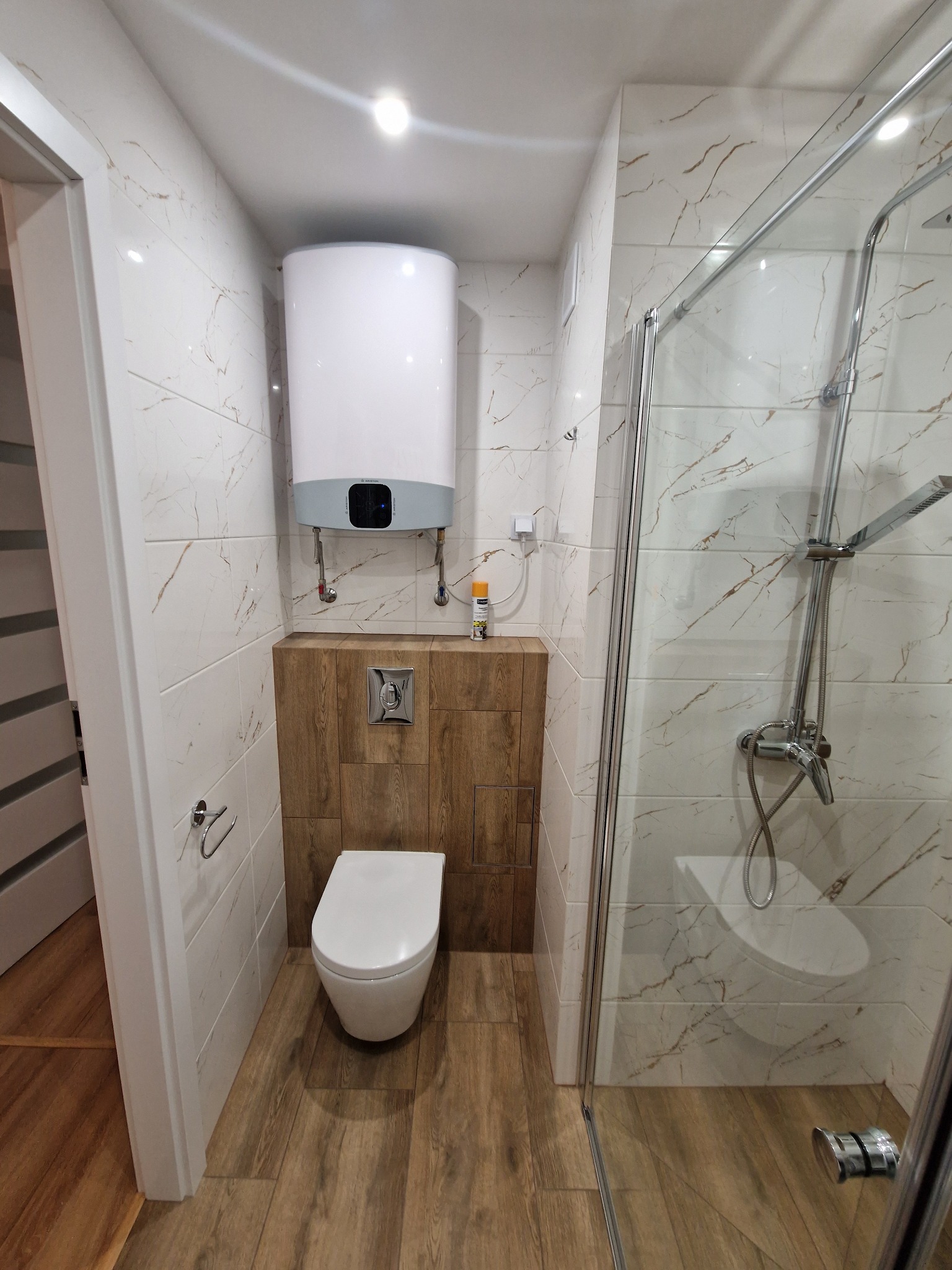 Nowoczesna łazienka po remoncie: białe płytki imitujące marmur, kabina prysznicowa ze szklanymi drzwiami, podwieszana toaleta z zabudową z drewnopodobnych paneli, bojler elektryczny na ścianie.