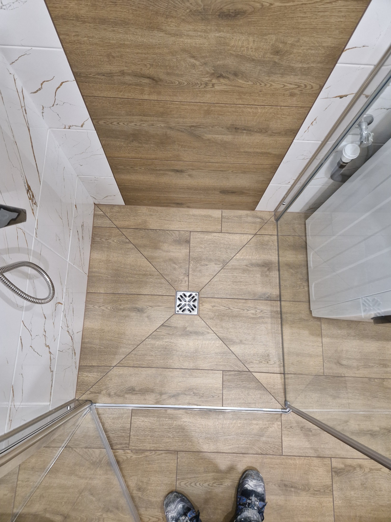 Widok z góry na wykończoną łazienkę z kabiną prysznicową, płytki podłogowe imitujące drewno ułożone w kwadrat wokół odpływu liniowego, białe płytki imitujące marmur na ścianach, w rogu widoczna...