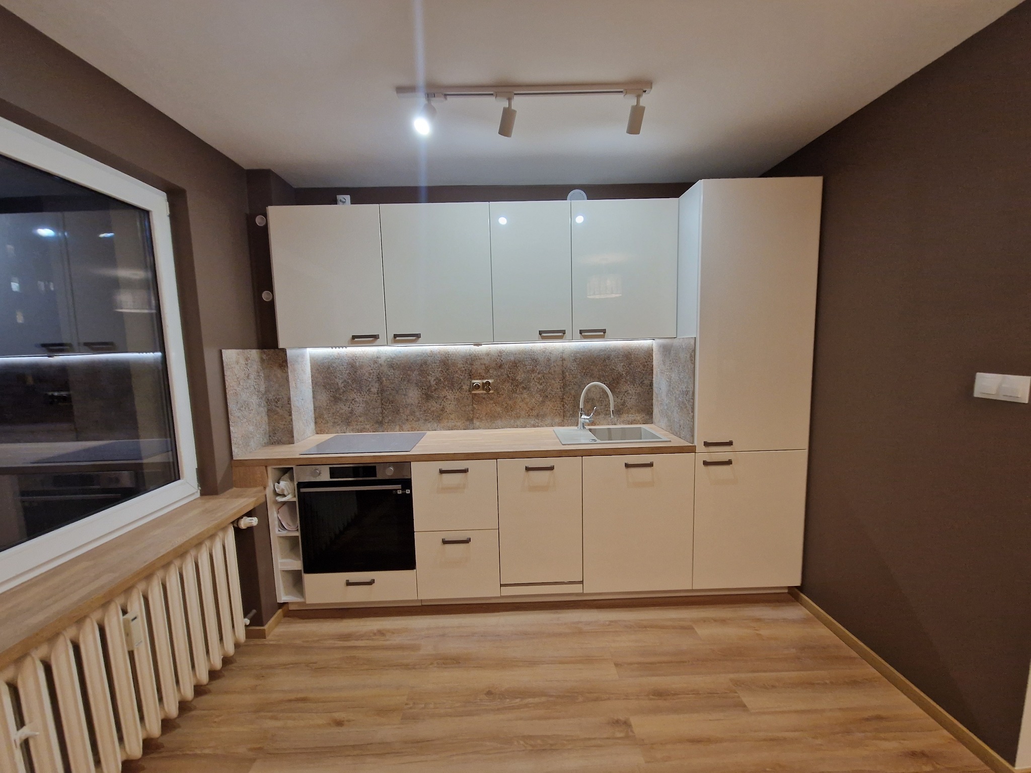 Nowoczesna, jasna kuchnia z białymi szafkami, drewnianym blatem i panelami podłogowymi w odcieniach brązu. Nad blatem zamontowane oświetlenie LED. Ściana pomalowana na ciemny brąz kontrastuje...