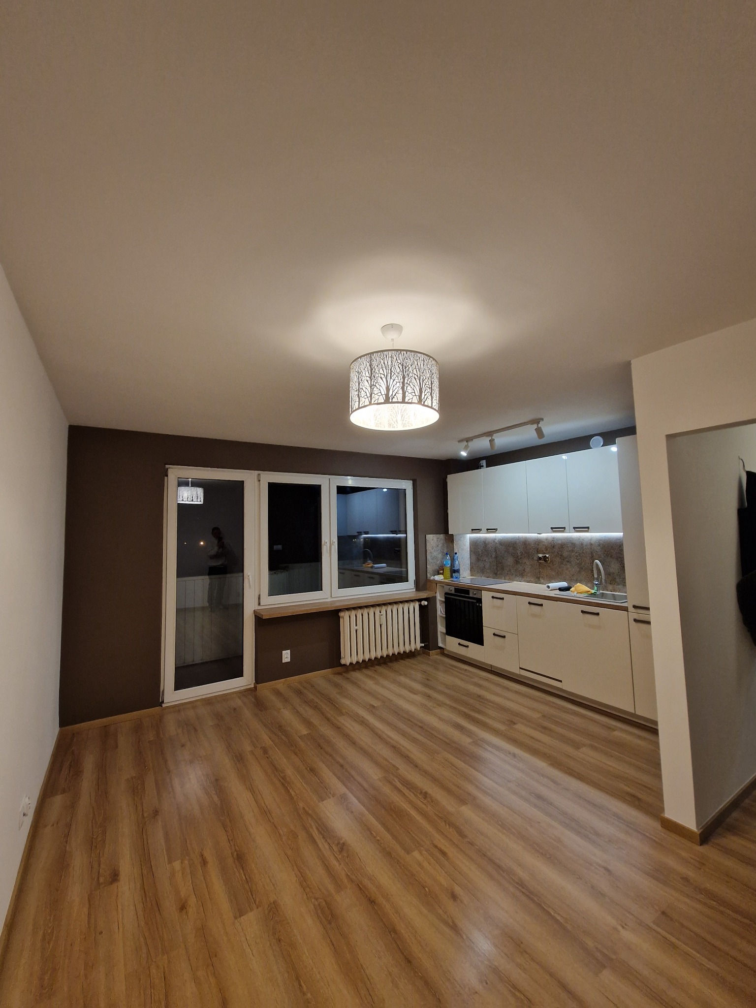 Nowoczesne wnętrze po remoncie: jasna kuchnia z białymi szafkami, drewnopodobna podłoga, brązowa ściana, okno balkonowe i dekoracyjna lampa sufitowa.