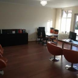 Jasne wnętrze biura z brązową podłogą, rzędem książek na szafce, dwoma biurkami z komputerami oraz krzesłami biurowymi i pomarańczowymi fotelami na chromowanych podstawach.