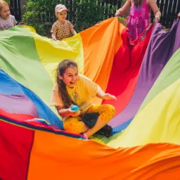 Uśmiechnięta dziewczynka siedzi na kolorowej chuście animacyjnej trzymanej przez inne dzieci i dorosłych, trzymając w dłoni niebieską piłkę.