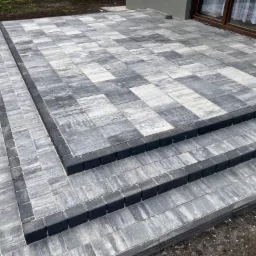 Schody i taras wyłożone szarą kostką o nieregularnym wzorze, obrzeża z ciemniejszej kostki, widok z góry.