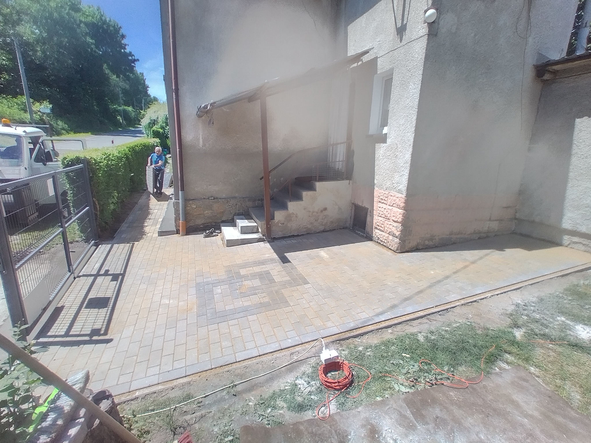 Ułożona kostka brukowa przed budynkiem z widocznym wejściem po schodach, obok furtka i pracownik z taczką w oddali, na pierwszym planie zwinięty przedłużacz.