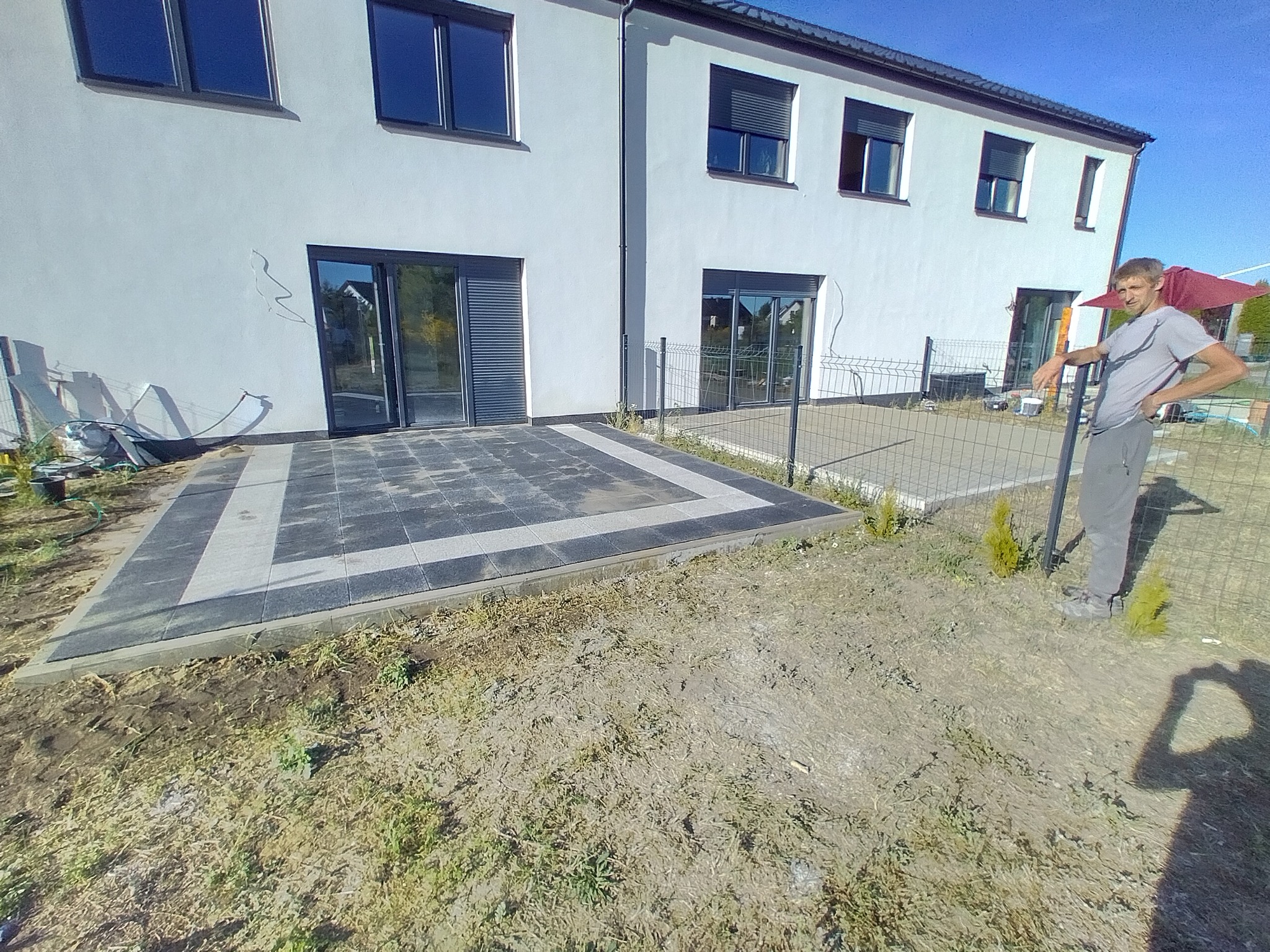 Wykończony taras z ciemnej i jasnej kostki na posesji nowego domu, widoczny pracownik opierający się o ogrodzenie z siatki.