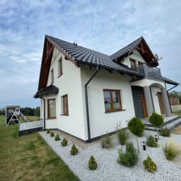 USŁUGI REMONTOWO-BUDOWLANE KACPER BORYS - Nowoczesny dom jednorodzinny z białymi ścianami, ciemną dachówką i drewnianymi elementami wykończeniowymi, z balkonem z metalową balustradą i zadbanym ogrodem z ozdobnymi krzewami i kamieniami,...