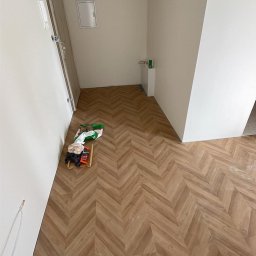 USŁUGI REMONTOWO-BUDOWLANE KACPER BORYS - Ułożona podłoga z paneli w jodełkę w jasnym wnętrzu, widoczne narzędzia i materiały budowlane w rogu pomieszczenia.