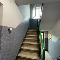 USŁUGI REMONTOWO-BUDOWLANE KACPER BORYS - Klatka schodowa w Mogilnie po malowaniu ścian na szaro z widocznymi schodami i zieloną barierką, otwarte okno na półpiętrze.