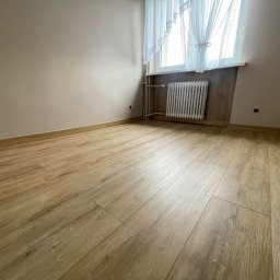 USŁUGI REMONTOWO-BUDOWLANE KACPER BORYS - Jasne panele podłogowe imitujące drewno ułożone w pustym pokoju z beżowymi ścianami, białym grzejnikiem pod oknem z koronkowymi firankami.