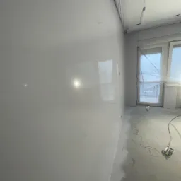 Świeżo położona, gładka, jasnoszara ściana w pokoju, z widocznym sprzętem do natryskowego nakładania gładzi i folią zabezpieczającą okno i sufit.