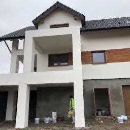 Nowoczesny dom jednorodzinny w trakcie malowania elewacji na biało, widoczne puste wiaderka po farbie, pracownik w jaskrawej kurtce i fragment nieotynkowanej ściany garażu.