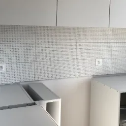 Biała kuchnia w trakcie montażu, widoczne płytki z drobnym wzorem na ścianie nad blatem i gniazdka elektryczne, szafki bez frontów.