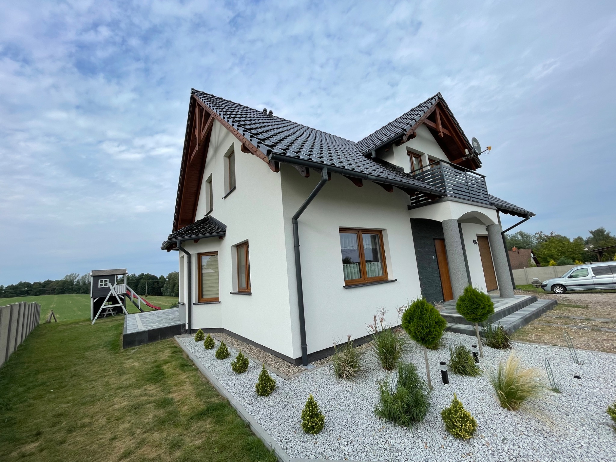 Nowoczesny dom jednorodzinny z białymi ścianami, ciemną dachówką i drewnianymi elementami wykończeniowymi, z balkonem z metalową balustradą i zadbanym ogrodem z ozdobnymi krzewami i kamieniami,...