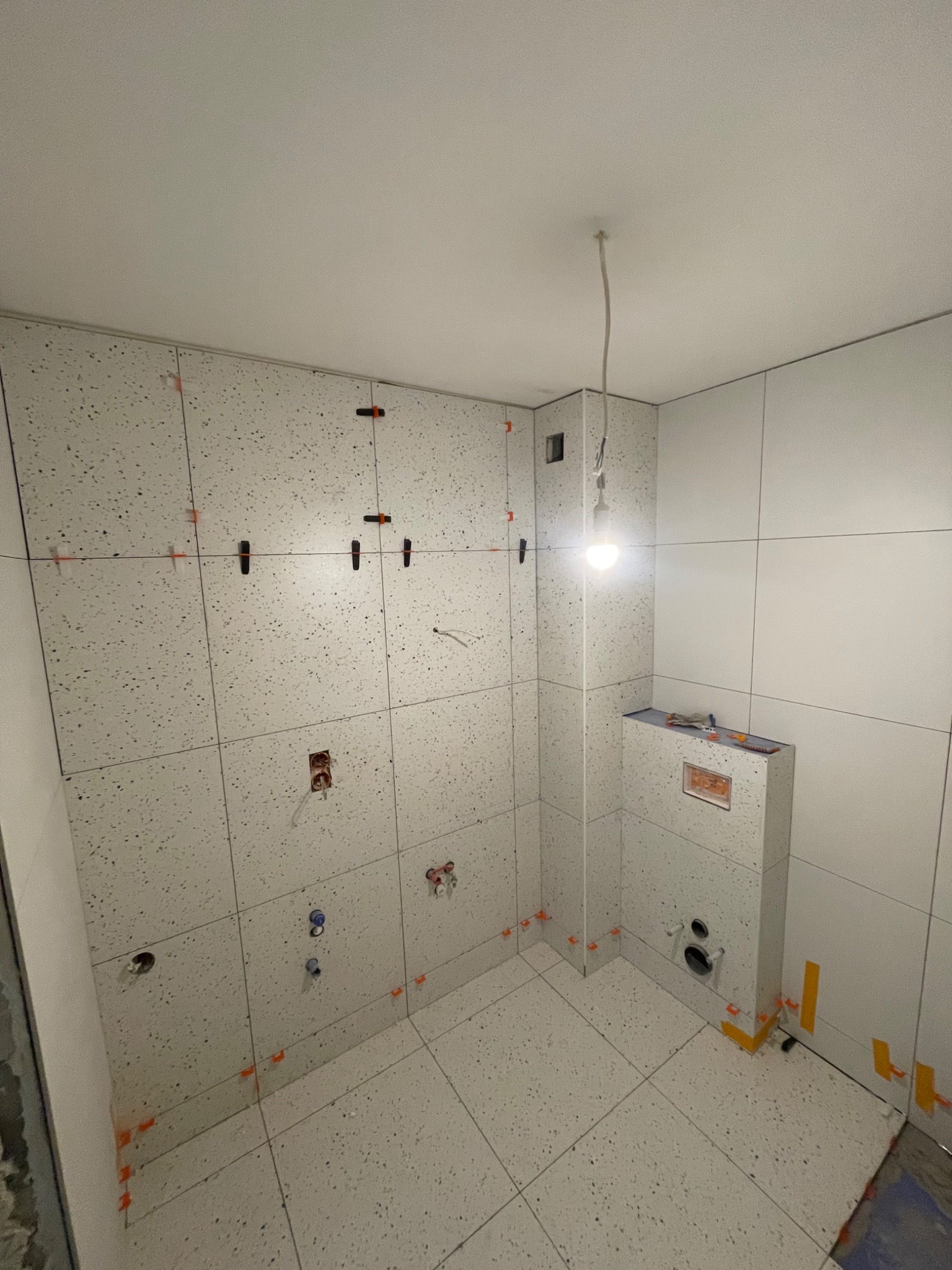 Białe płytki z wzorem lastryko układane w łazience, widoczne krzyżyki dystansowe i wystające przewody instalacji elektrycznej i wodnej, surowa żarówka oświetla pomieszczenie.