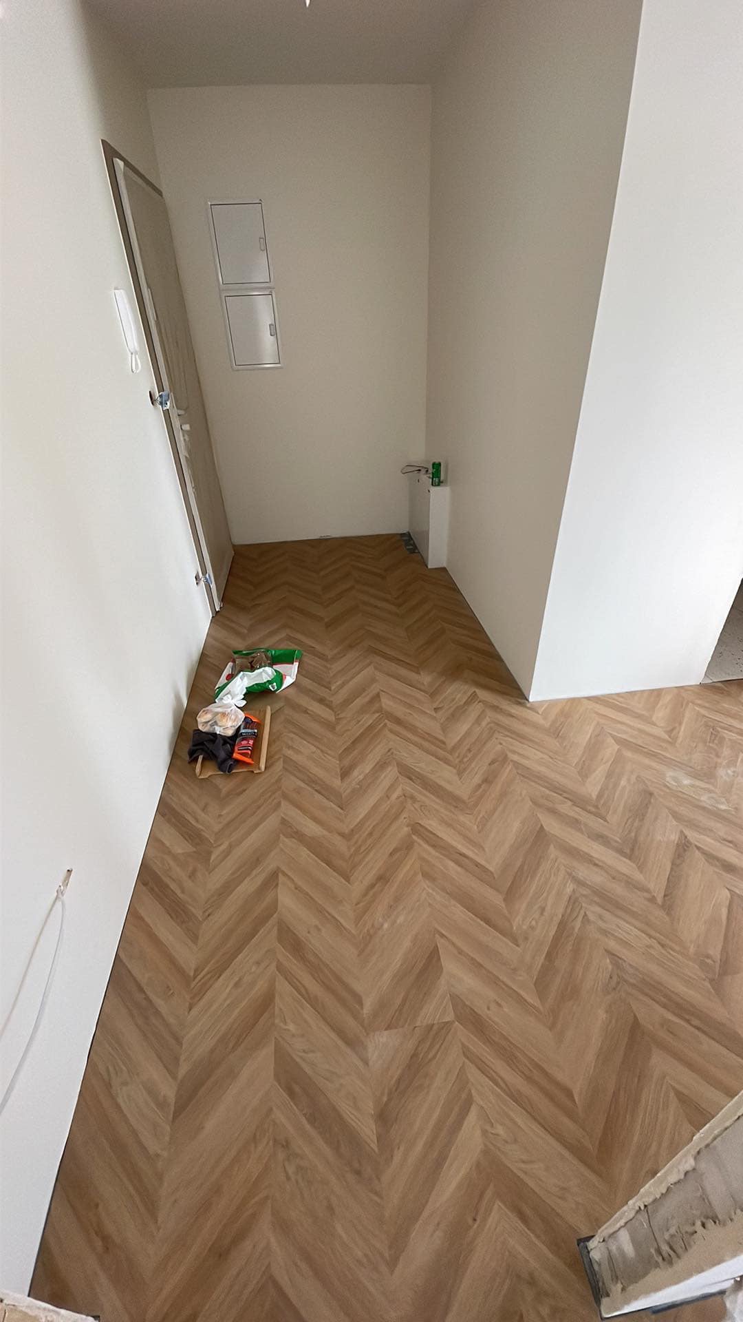 Ułożona podłoga z paneli w jodełkę w jasnym wnętrzu, widoczne narzędzia i materiały budowlane w rogu pomieszczenia.