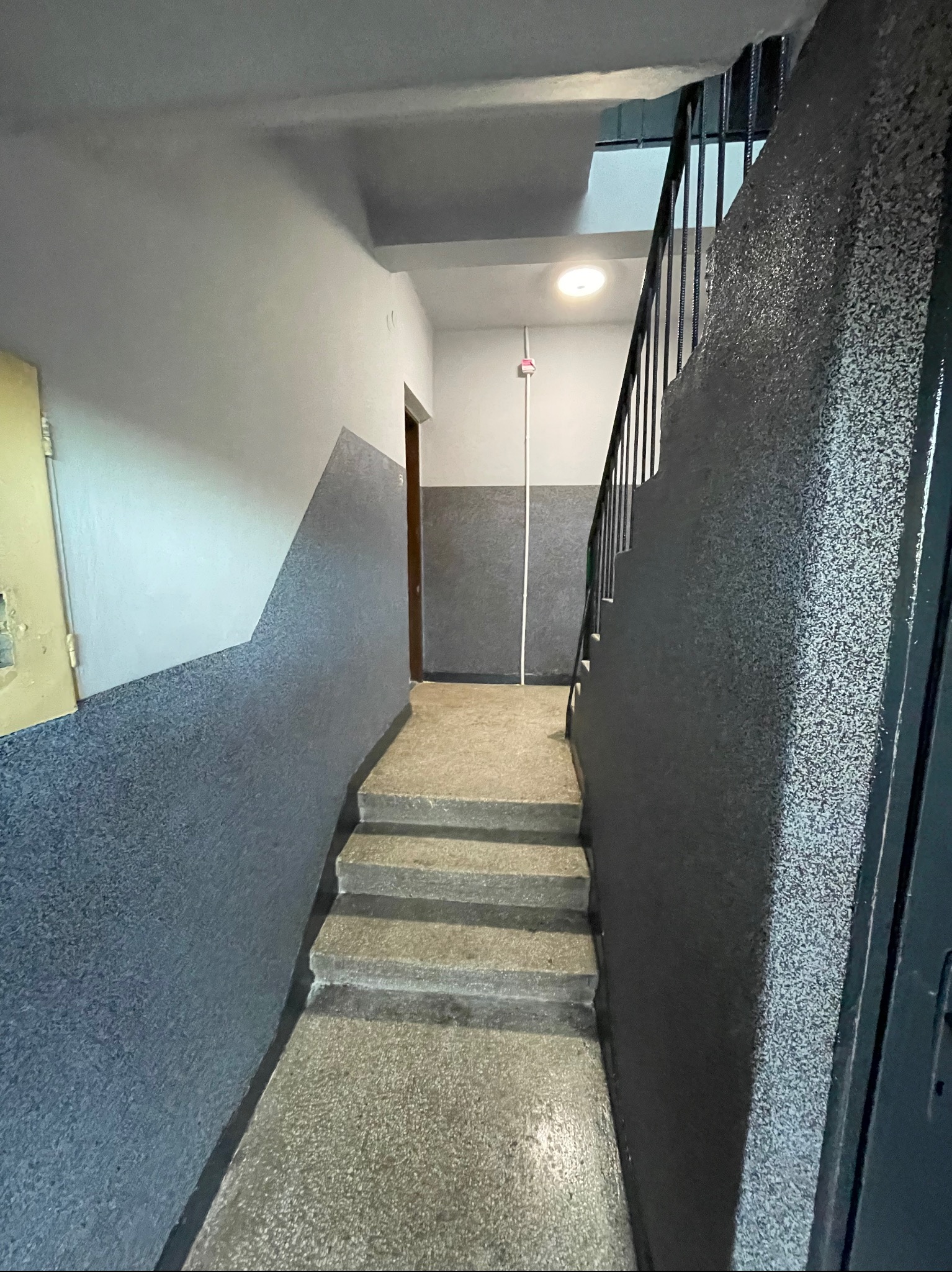 Klatka schodowa w bloku z szarymi ścianami i schodami, widoczne ślady użytkowania, charakterystyczny podział kolorystyczny ścian na dwie strefy, oświetlenie sufitowe, metalowa barierka schodowa.