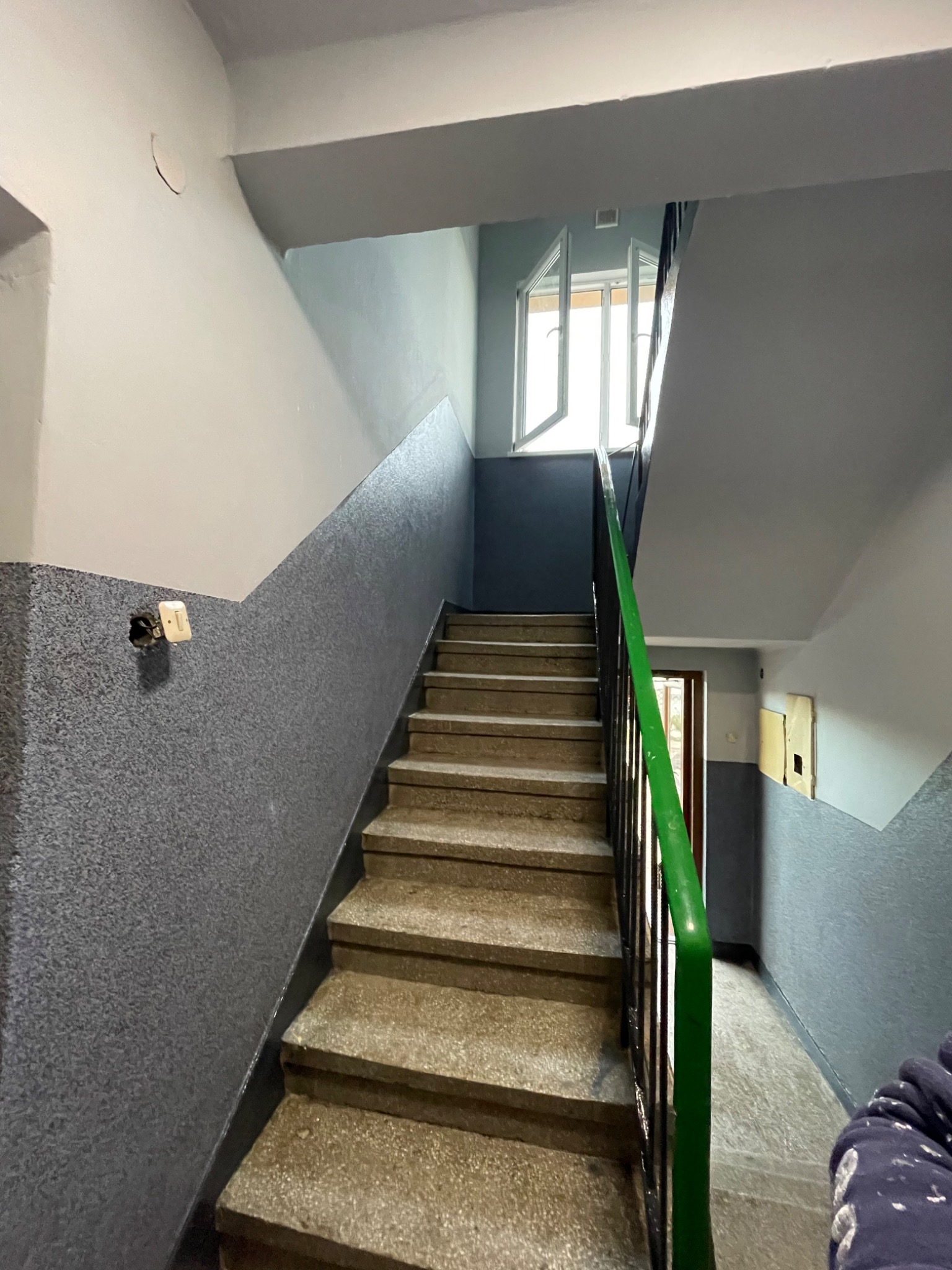 Klatka schodowa w Mogilnie po malowaniu ścian na szaro z widocznymi schodami i zieloną barierką, otwarte okno na półpiętrze.