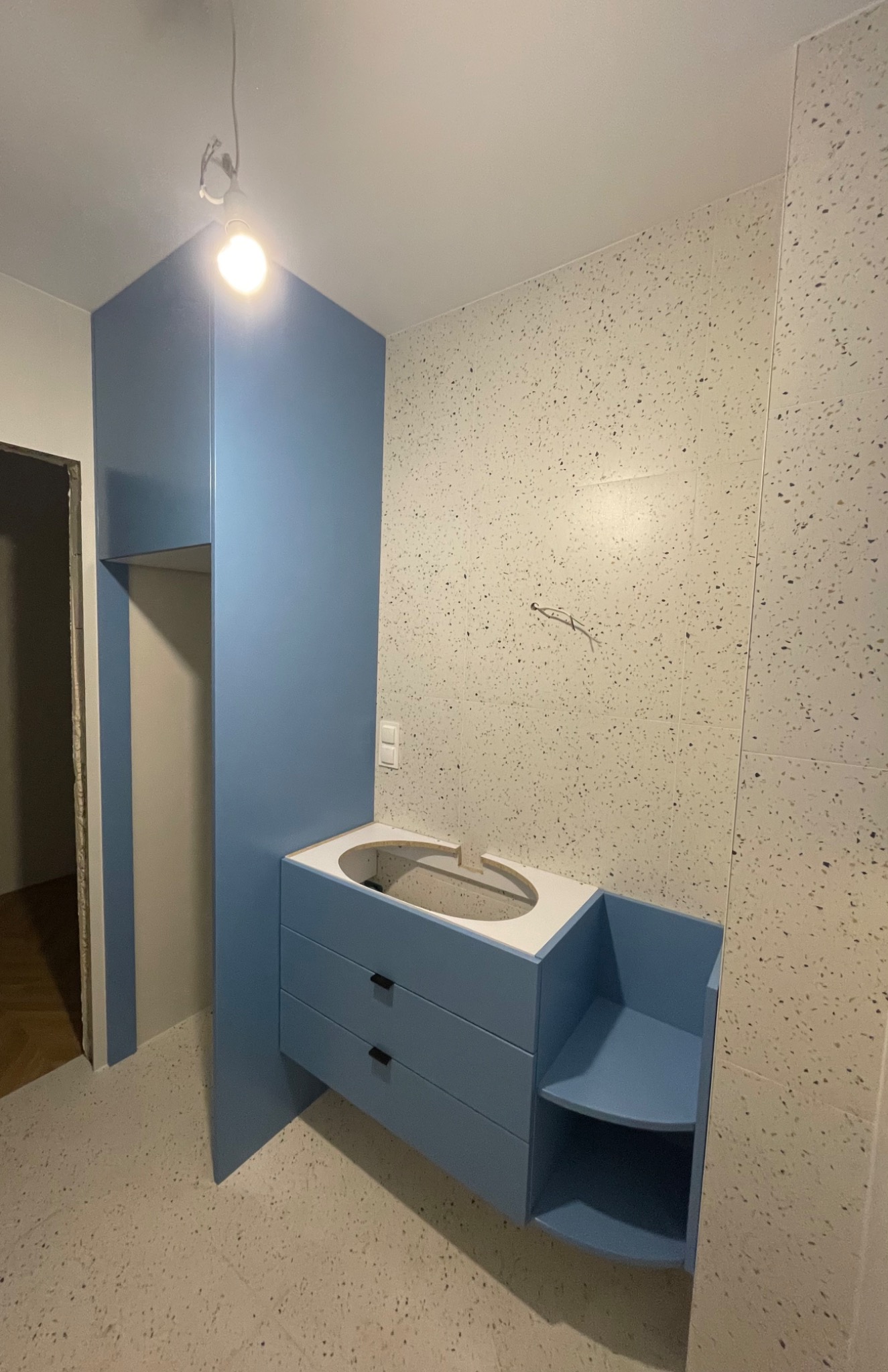 Niedokończona łazienka z niebieską szafką pod umywalkę z szufladami i półkami, ściany i podłoga wyłożone płytkami w stylu lastryko, widoczne wystające kable elektryczne.