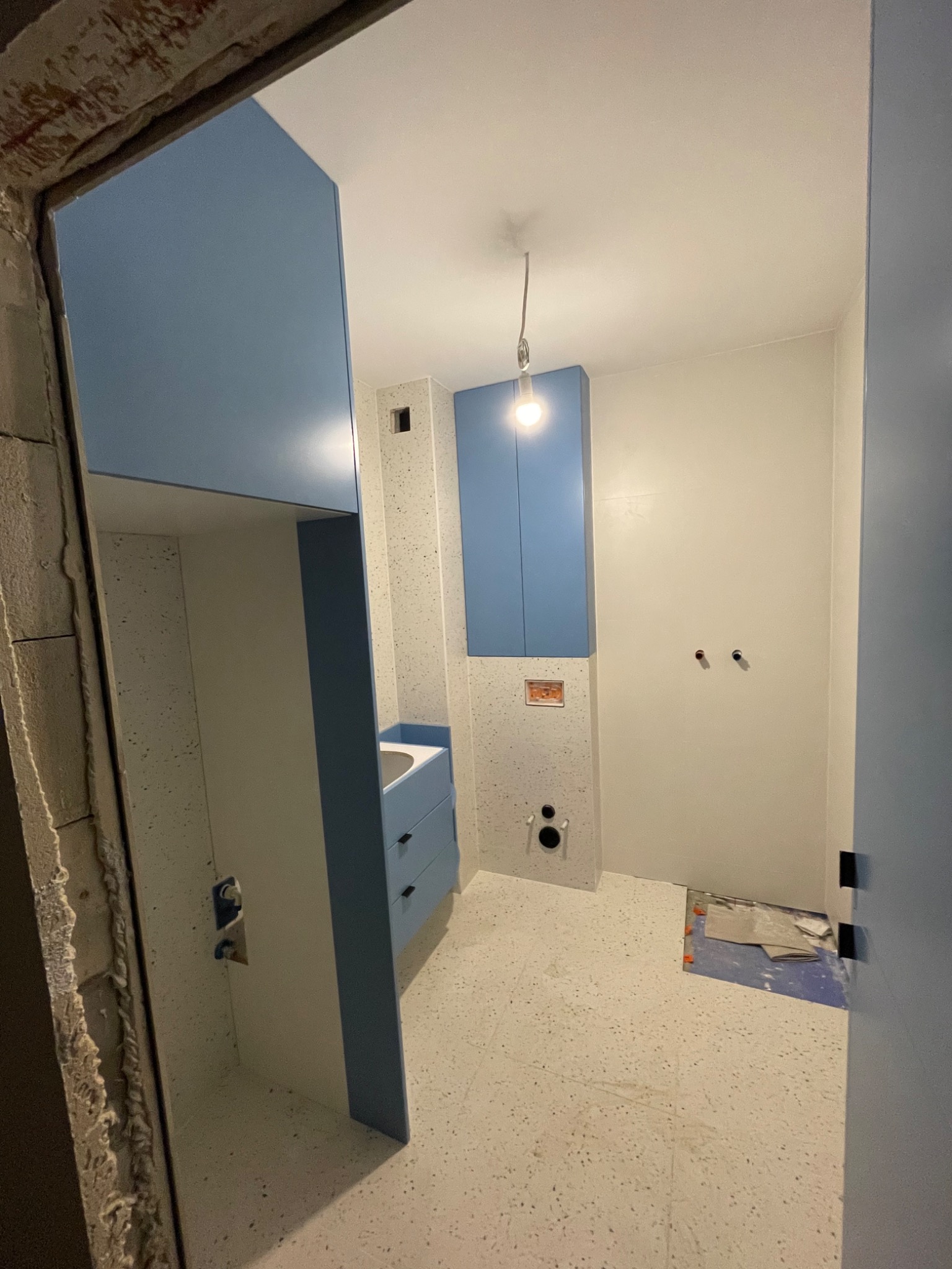 Łazienka w trakcie remontu, widoczne niebieskie szafki, białe ściany, terazzo na ścianach i podłodze, surowy ościeżnica drzwi i niedokończona podłoga.