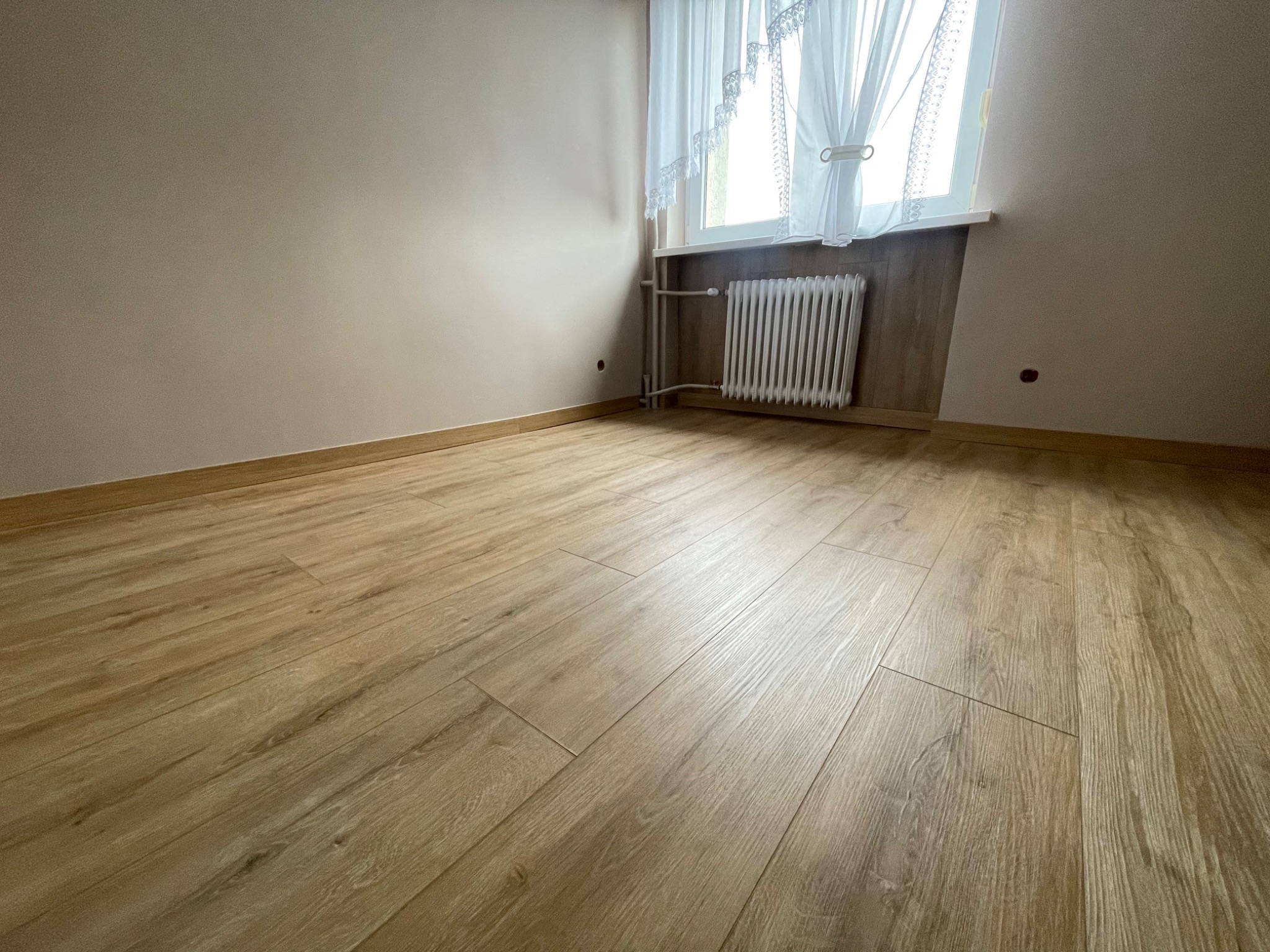 Jasne panele podłogowe imitujące drewno ułożone w pustym pokoju z beżowymi ścianami, białym grzejnikiem pod oknem z koronkowymi firankami.