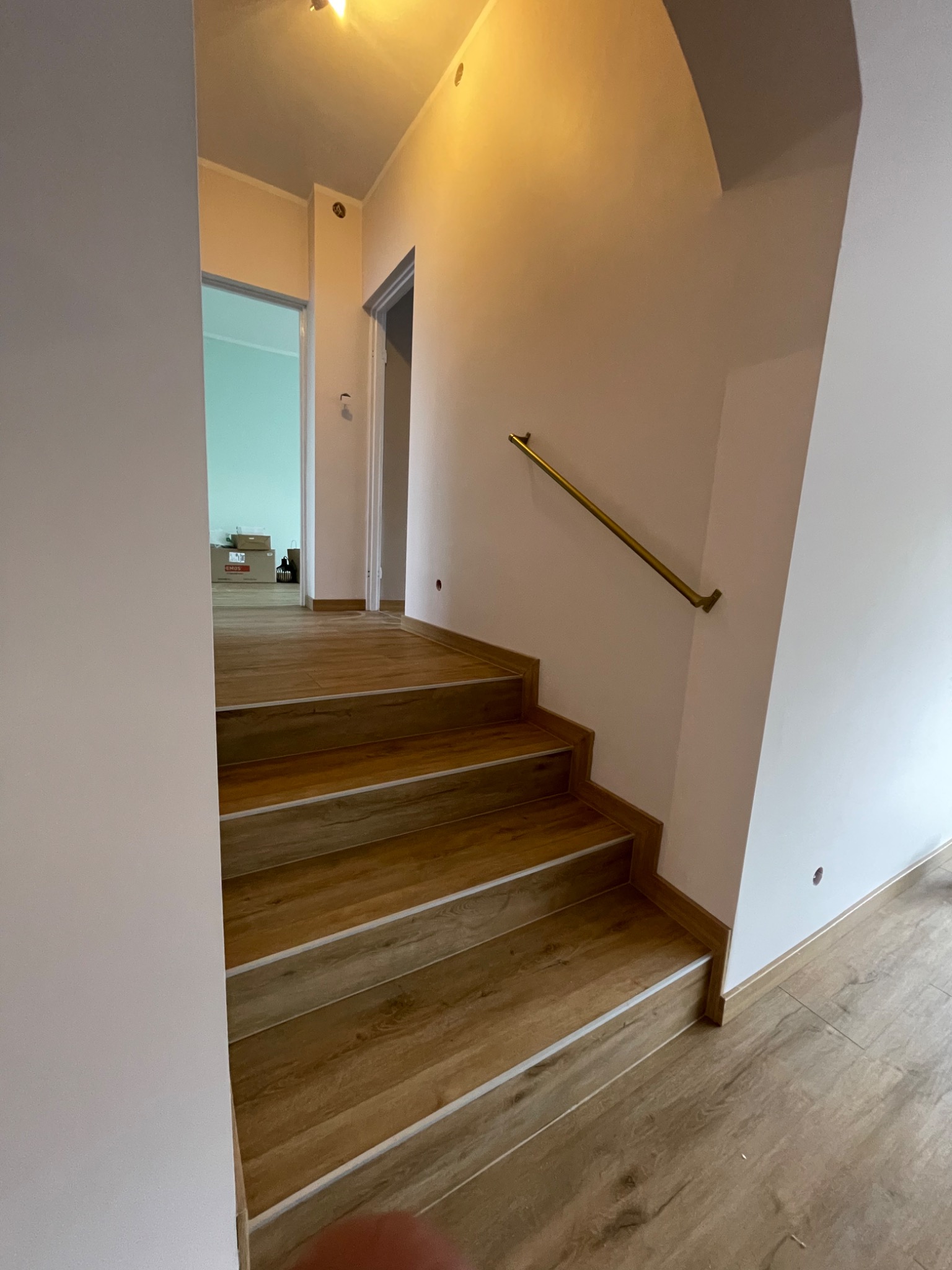 Schody wyłożone panelami imitującymi drewno, z zamontowaną złotą poręczą, widok z dołu schodów na otwarte drzwi do innego pomieszczenia.