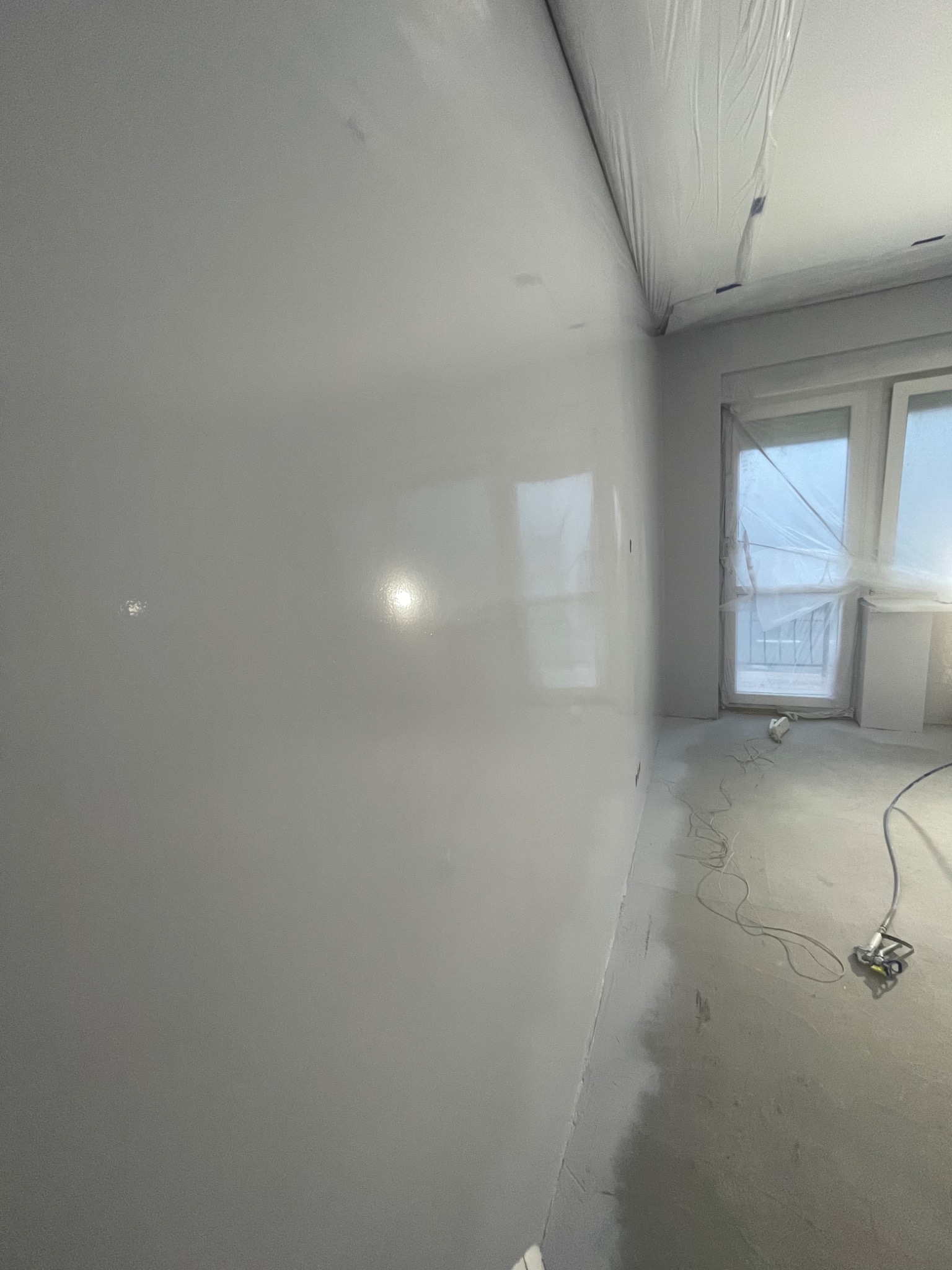 Świeżo położona, gładka, jasnoszara ściana w pokoju, z widocznym sprzętem do natryskowego nakładania gładzi i folią zabezpieczającą okno i sufit.