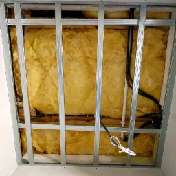 Ocieplenie poddasza wełną mineralną widoczne przez metalowy stelaż konstrukcji sufitu podwieszanego, z widocznymi przewodami elektrycznymi.