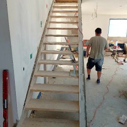 Ardes - Wnętrze w trakcie budowy z widocznymi drewnianymi schodami w surowym stanie, poziomica przy ścianie i pracownik w tle, kable wystające ze ściany.