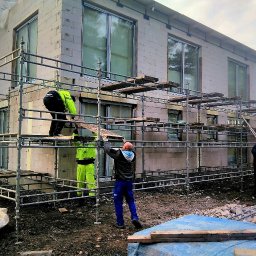 Ardes - Budowa domu z białych bloczków: robotnicy na rusztowaniu montują elementy konstrukcyjne, widoczne duże okna i surowy krajobraz.