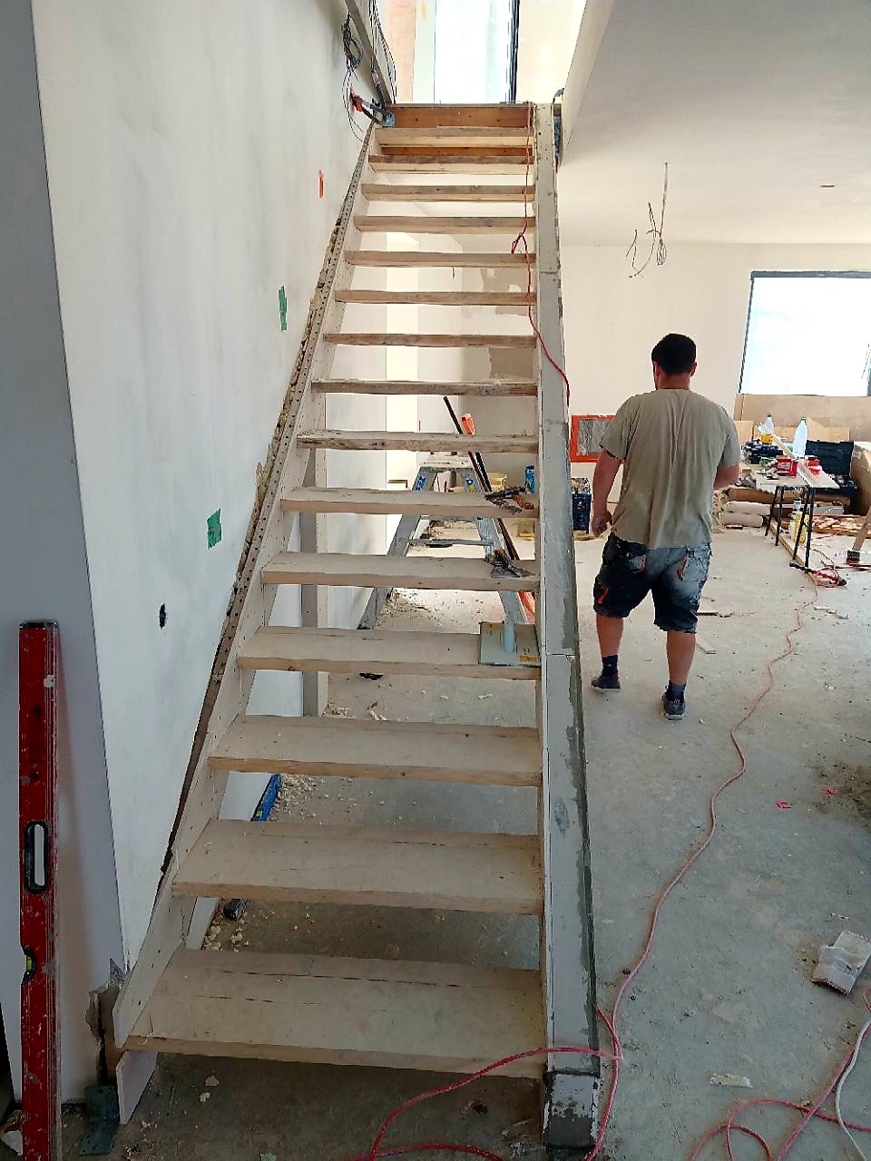 Wnętrze w trakcie budowy z widocznymi drewnianymi schodami w surowym stanie, poziomica przy ścianie i pracownik w tle, kable wystające ze ściany.
